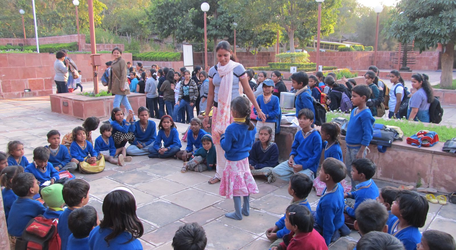 Children seated on the museum forecourt in groups with their teachers, the meal container standing among them.