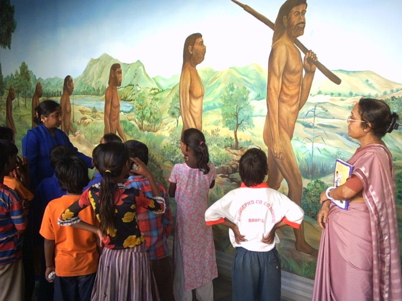 A teacher and a group of children standing in front of a painted mural of human evolution inside the Regional Museum of Natural History.